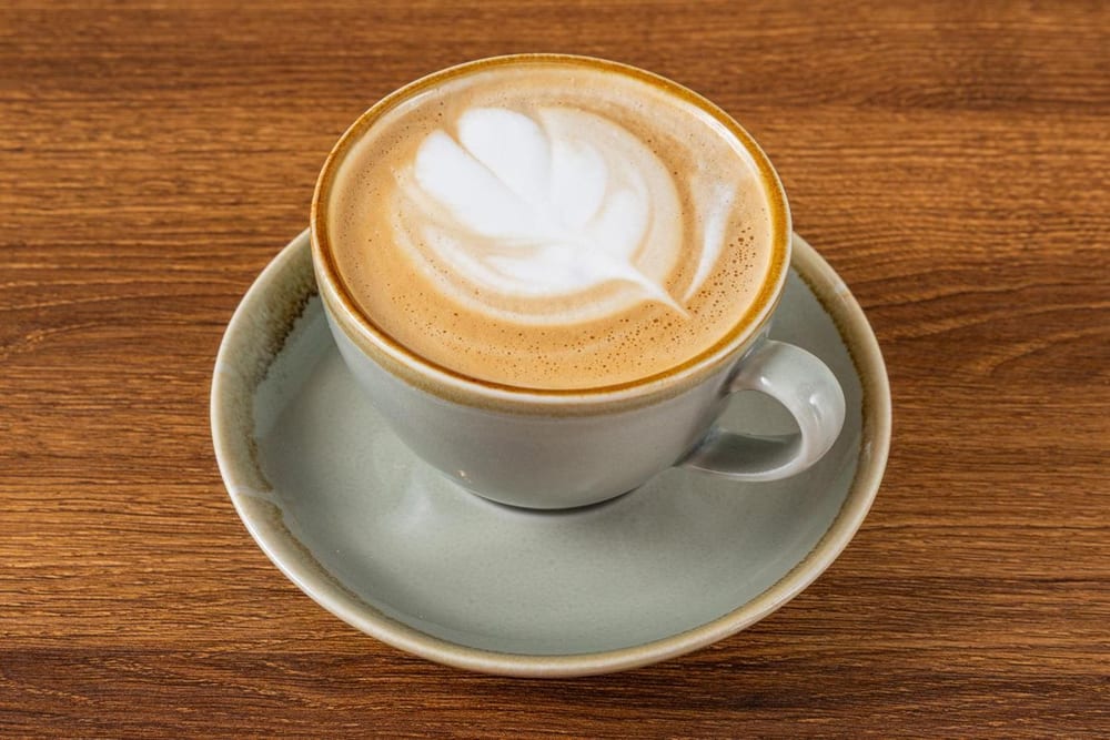 A latte with a rosetta of milk foam in a sage-green cup and saucer on a wooden table