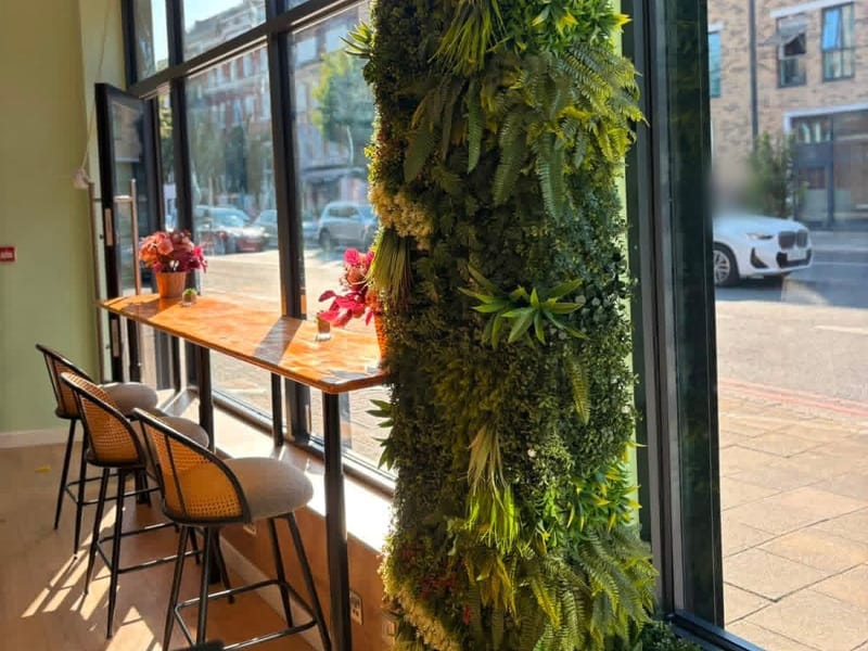 A wall of greenery beside floor-to-ceiling windows, with rattan stools and flowers along a wooden window counter