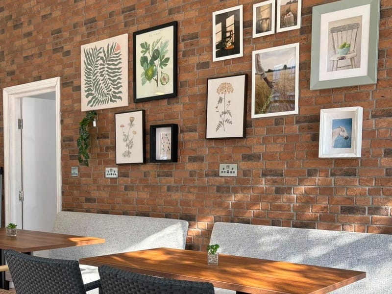 Bench seating along an exposed brick wall hung with botanical prints and framed photographs, with wicker chairs at a sunlit wooden table